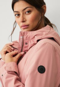 Chaqueta impermeable rosa con capucha, que cuenta con un zipper frontal y botones a presión. Incluye un parche redondo oscuro en la manga izquierda como detalle.