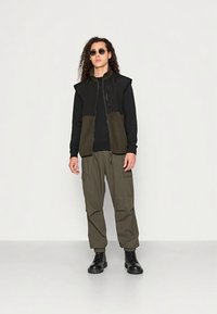 Junge Person mit langen lockigen Haaren, die Sonnenbrillen trägt, eine schwarze und olivfarbene Weste, schwarze Langarmshirts, olivfarbene Cargo-Hosen und schwarze Stiefel, die an einer weißen Wand steht.