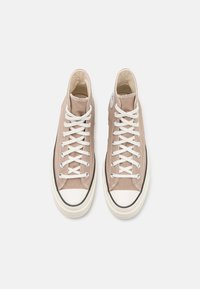 Baskets montantes en toile brune clair avec des lacets blancs, une coque en caoutchouc crème à l'avant et un contour de semelle noire. Présente une patch logo circulaire sur le côté.