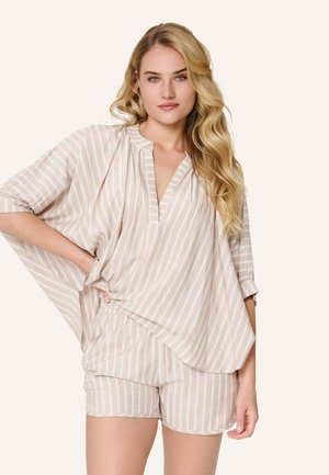 Donna bionda con capelli ondulati, che indossa un top largo a righe verticali beige e bianche e pantaloncini coordinati, in piedi con una mano sui fianchi.