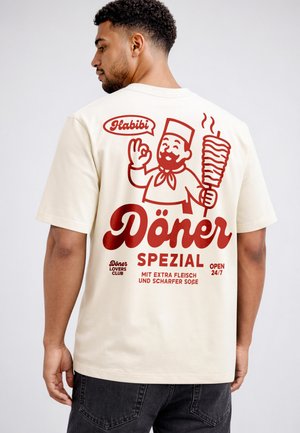 Man wearing cream t-shirt with red döner graphic, chef illustration holding meat skewer, and text in German about extra meat and spicy sauce.
