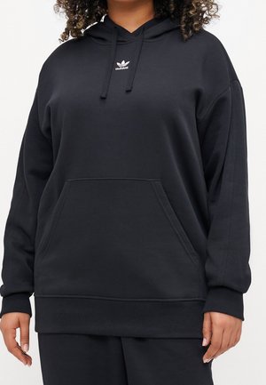 Personne portant un sweat à capuche Adidas noir avec une poche avant et des cordons, montrant le haut du corps et les bras.