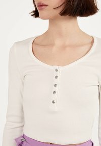 Femme portant un crop top blanc côtelé ajusté avec cinq boutons argentés sur le devant, associé à un pantalon violet taille haute.