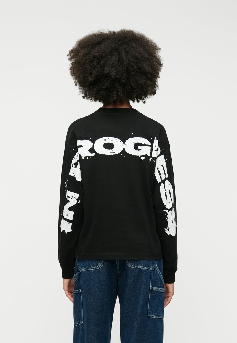 Melna long-sleeve T-krekla ar lieliem baltiem uzrakstiem uz muguras, atbrīvota piegriezuma un ribinātu kakla izgriezumu. Papildināti ar džinsu cargo biksēm.