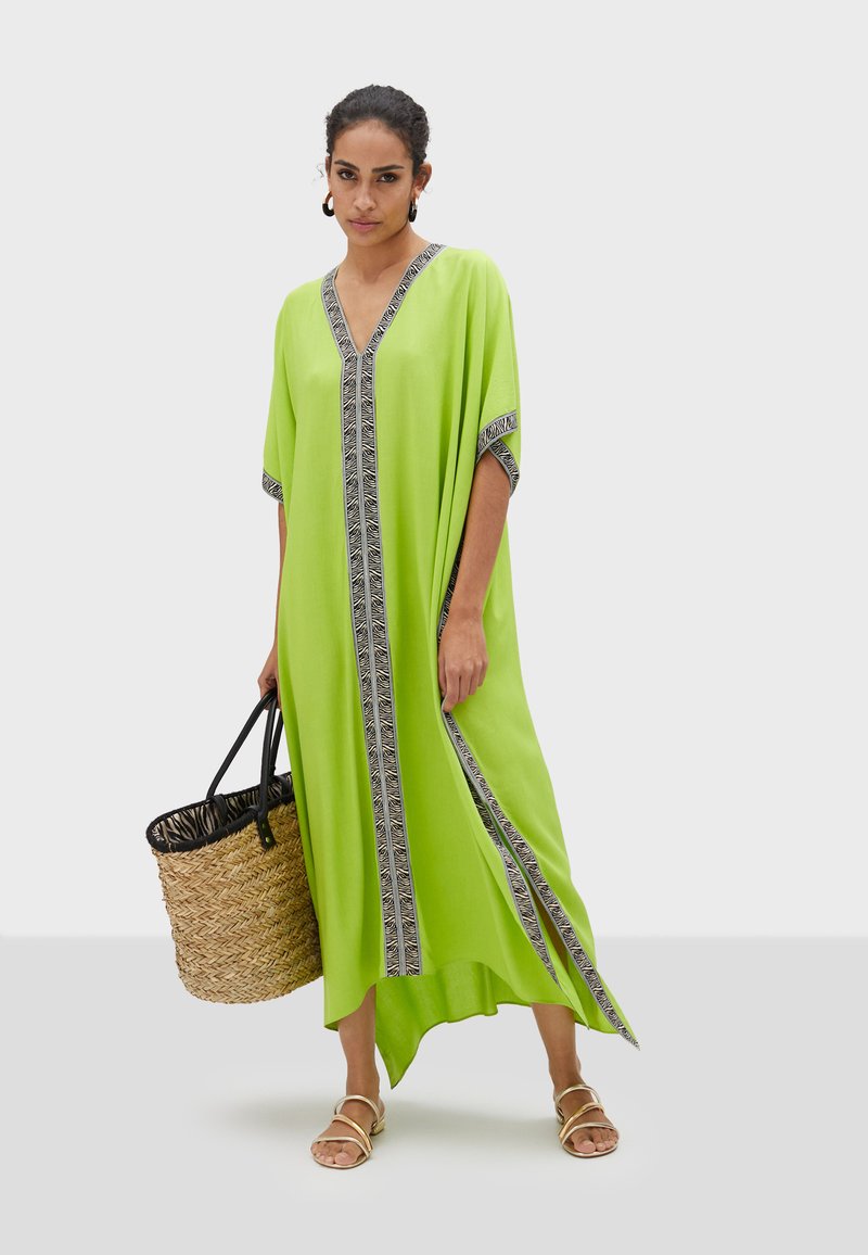 Limegrön kaftan med V-ringning, korta ärmar och kontrasterande svart och vit mönstrad kant längs fram. Modell håller en vävd väska.