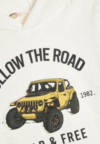 Witte hoodie met een gele geïllustreerde jeep graphic, inclusief zwarte tekst "VOLG DE WEG" en "1982." De tekst is vetgedrukt en opvallend.