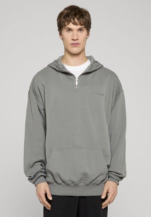 Fiatal férfi laza, szürke félcipzáras kapucnis pulóverben, első zsebbel és fehér aláöltözővel, egyszerű világos háttér előtt áll.