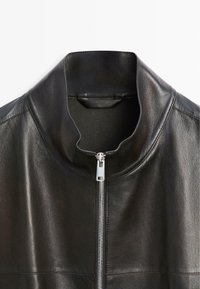 Zwart leren jack met een opstaande kraag en een zilveren rits aan de voorkant, gezien vanaf de bovenkant vooraan tegen een witte achtergrond.