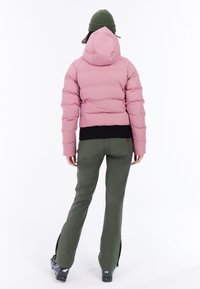 Růžová péřová bunda s kapucí, elastický černý lem, doplněná přiléhavými zelenými lyžařskými kalhotami. Viditelné zipy na kotnících kalhot a černé lyžařské boty.