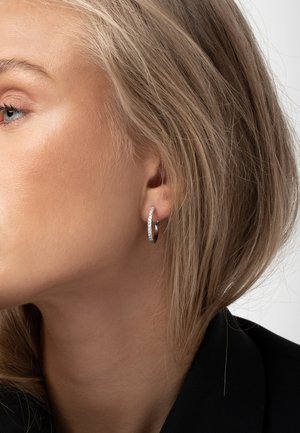 Blond kvinde med kort hår iført en sølv ørering med små indlejrede sten, vendt mod venstre på en ensfarvet baggrund.