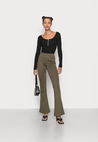 Top preto de mangas compridas com um detalhe em zip, combinado com calças flare verdes-oliva. O modelo segura uma pequena mala preta; os sapatos apresentam um design volumoso.