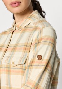 Camicia a quadri beige con accenti arancioni, blu e gialli. Presenta un colletto con bottoni e una tasca sul petto. Include un'etichetta con logo marrone sulla manica.
