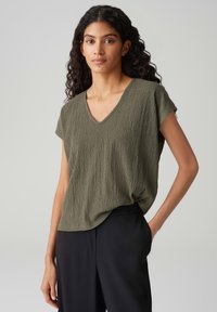Olive green, textured V-neck top with short sleeves, draped fit, paired with black trousers. Simple design, relaxed silhouette.