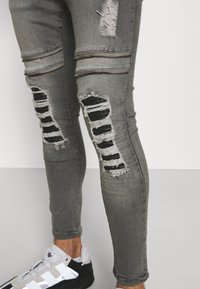 Jeans gris de corte slim con detalles desgastados, cremalleras horizontales y acentos de tela negra en las áreas rasgadas. Presentan un dobladillo deshilachado y una tela texturizada.