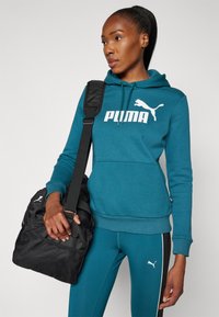 Felpa teal con logo "PUMA" bianco, tasca a marsupio e stringhe. Borsa sportiva nera con dettaglio in rete tenuta in mano. Leggings aderenti con strisce.