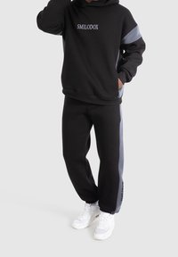 Schwarzer Hoodie mit grauen Akzenten, versehen mit gesticktem "SMILODOX", kombiniert mit passenden schwarzen Hosen und weißen Sneakers.