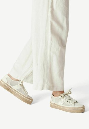 Pieds portant des baskets plateformes blanc cassé avec un détail en corde tressée, associés à un pantalon large, fluide et clair.