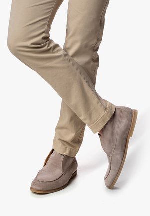 Zapatos de ante gris con un perfil bajo y lados abiertos, combinados con pantalones beige claros. Textura suave, suela plana y detalles de costura mínimos.