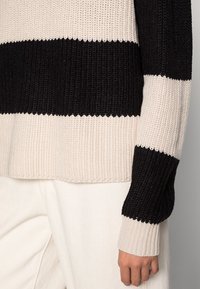 Schwarz-beiger gestreifter Strickpullover mit gerippter Textur. Der Ärmel hat einen kontrastierenden schwarzen Abschnitt, der die Strickdetails und die Struktur hervorhebt.