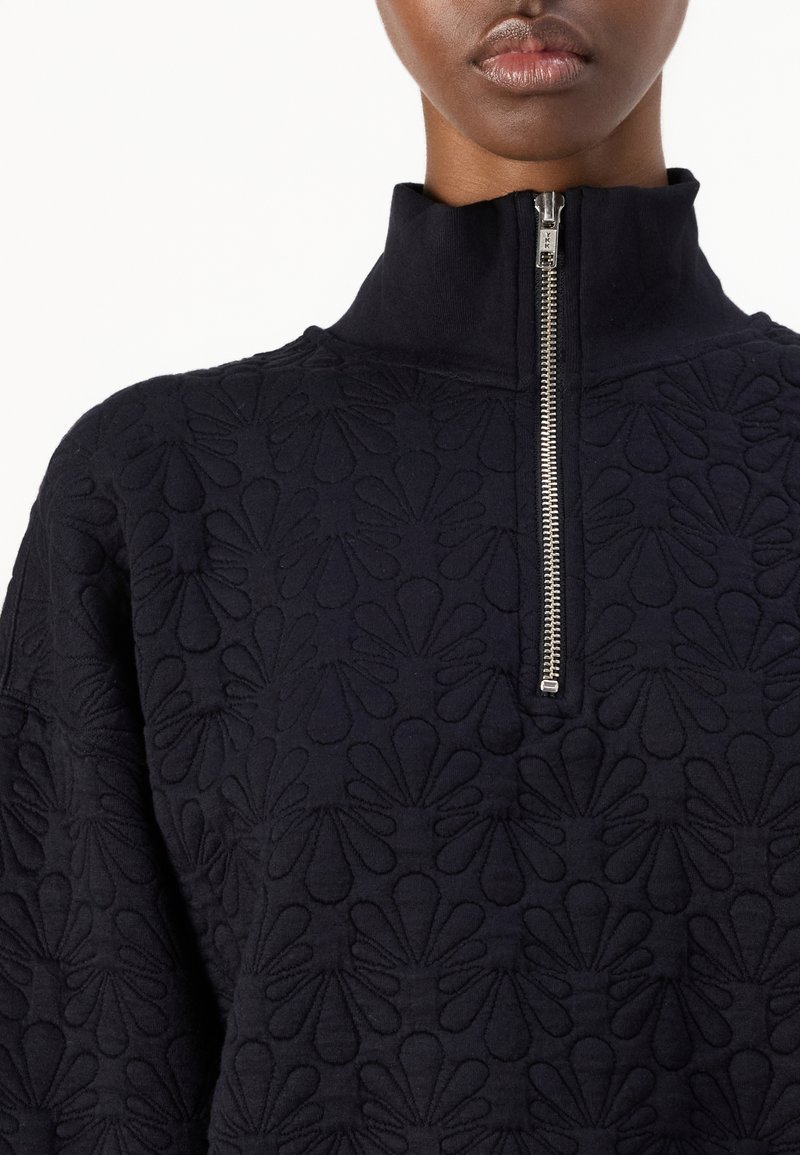 Top negro de cuello alto con un patrón floral texturizado, cierre frontal de media cremallera y tejido suave con un sutil brillo.