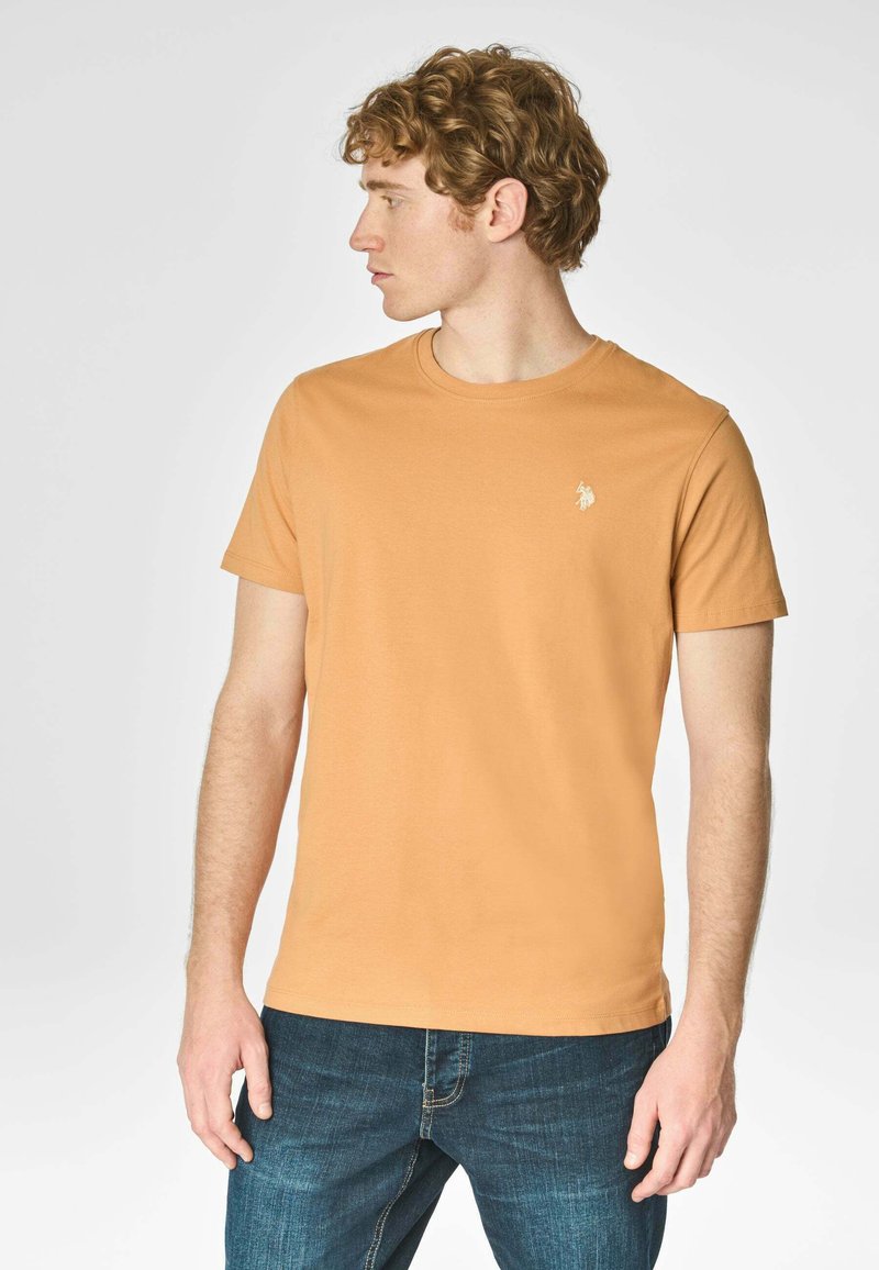 T-shirt a maniche corte con scollatura rotonda di colore arancione tenue. Realizzato in un tessuto morbido, presenta un piccolo logo bianco sul petto. Abbinato a jeans blu.