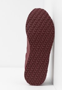 Semelle d'une chaussure bordeaux présentant un motif de bande de roulement texturé en zigzag, fabriquée en caoutchouc. Le côté est partiellement visible.