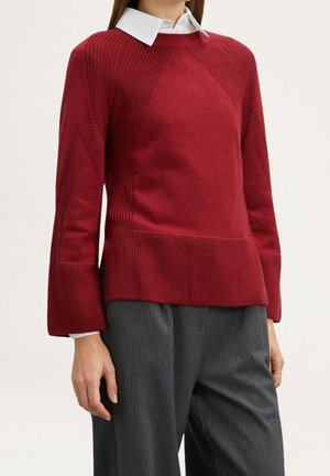 Mujer vistiendo un suéter de punto rojo con textura sobre una camisa blanca con cuello, combinado con pantalones de pierna ancha gris con rayas diplomáticas.