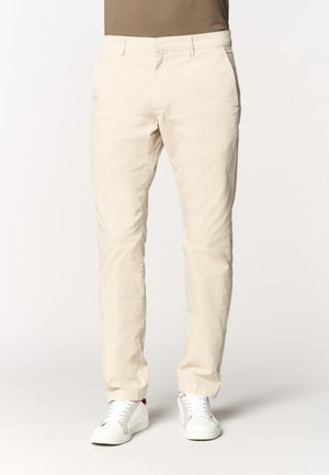 Pantaloni chinos beige slim fit da uomo abbinati a sneakers bianche, mostrati dalla vita ai piedi su uno sfondo neutro.