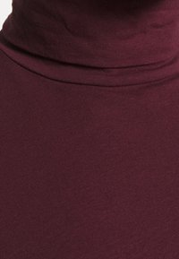 Bordowy golf wykonany z miękkiej, teksturowanej tkaniny o dopasowanym kroju. Posiada wysoki kołnierz i gładką powierzchnię.