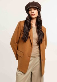 Donna con lunghi capelli scuri che indossa un cappello da autista marrone, un blazer color cammello, un top lavorato beige e pantaloni a palazzo color beige chiaro su uno sfondo semplice.