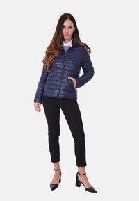 Giacca puffer blu navy con chiusura a zip, texture trapuntata e tasche laterali, indossata con una camicia chiara, pantaloni neri e scarpe con tacco nere.