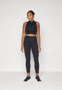 Top sportivo nero corto con zip frontale, abbinato a leggings neri a vita alta. Il modello indossa scarpe da ginnastica nere. Texture liscia.