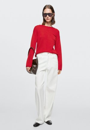 Donna con maglione rosso e pantaloni bianchi, che porta una borsa marrone e indossa occhiali da sole scuri, in piedi su uno sfondo chiaro e uniforme.