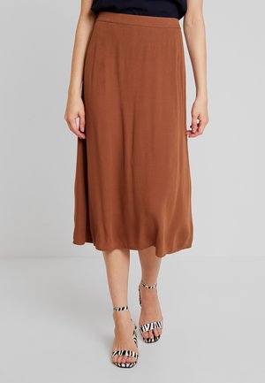 Woman wearing a brown midi skirt with a black top and zebra-striped ankle-strap sandals, standing on a gray floor against a gray background.