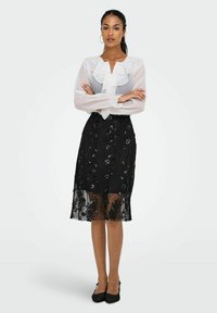 Bílá průhledná blůza s volánkovými detaily, v kombinaci s černou krajkovou sukní zdobenou flitry a květinovými vzory, celý outfit doplněný černými podpatky.