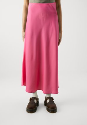 Kvinde iført en lang, lyserød nederdel, hvide sokker og brune sko med spænder, stående mod en ensfarvet hvid baggrund.