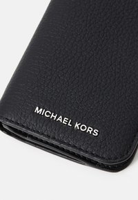 Svart lærpung med teksturert overflate og sølvfarget "MICHAEL KORS"-logo. Rektangulær form med avrundede kanter og glatt innside.