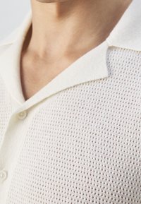 Polo shirt bianco a maniche corte realizzato in tessuto a rete, con colletto morbido, chiusura con bottoni e un motivo strutturato su tutta la superficie.