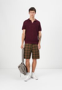 Polo shirt texturé bordeaux à manches courtes, associé à un short à carreaux marron. Le modèle porte un sac à bandoulière gris et des baskets montantes blanches.