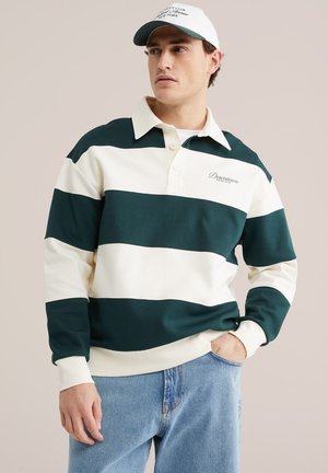 Mladý muž oblečený v bielom a tmavozelenom široko pruhovanom dlhom rukáve rugby trička, svetlomodrých džínsoch a bielej čiapke, stojaci s jednou rukou v vrecku.