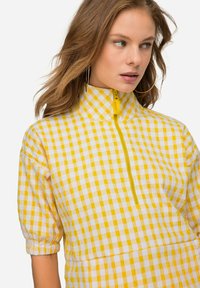 Camicia a quadri bianchi e gialli con collare alto, maniche a sbuffo corte e zip sul davanti. Tessuto strutturato con vestibilità comoda.