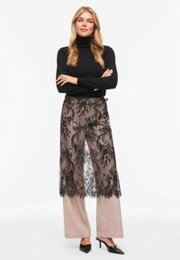 Overlay en dentelle noire avec motifs floraux sur tissu beige transparent, silhouette à jambes larges, assortie à un col roulé noir et des talons noirs.