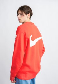 Crvena pletiva majica sa bijelim Nike logom na leđima, s okruglim izrezom i rebrastim manžetama i rubom.
