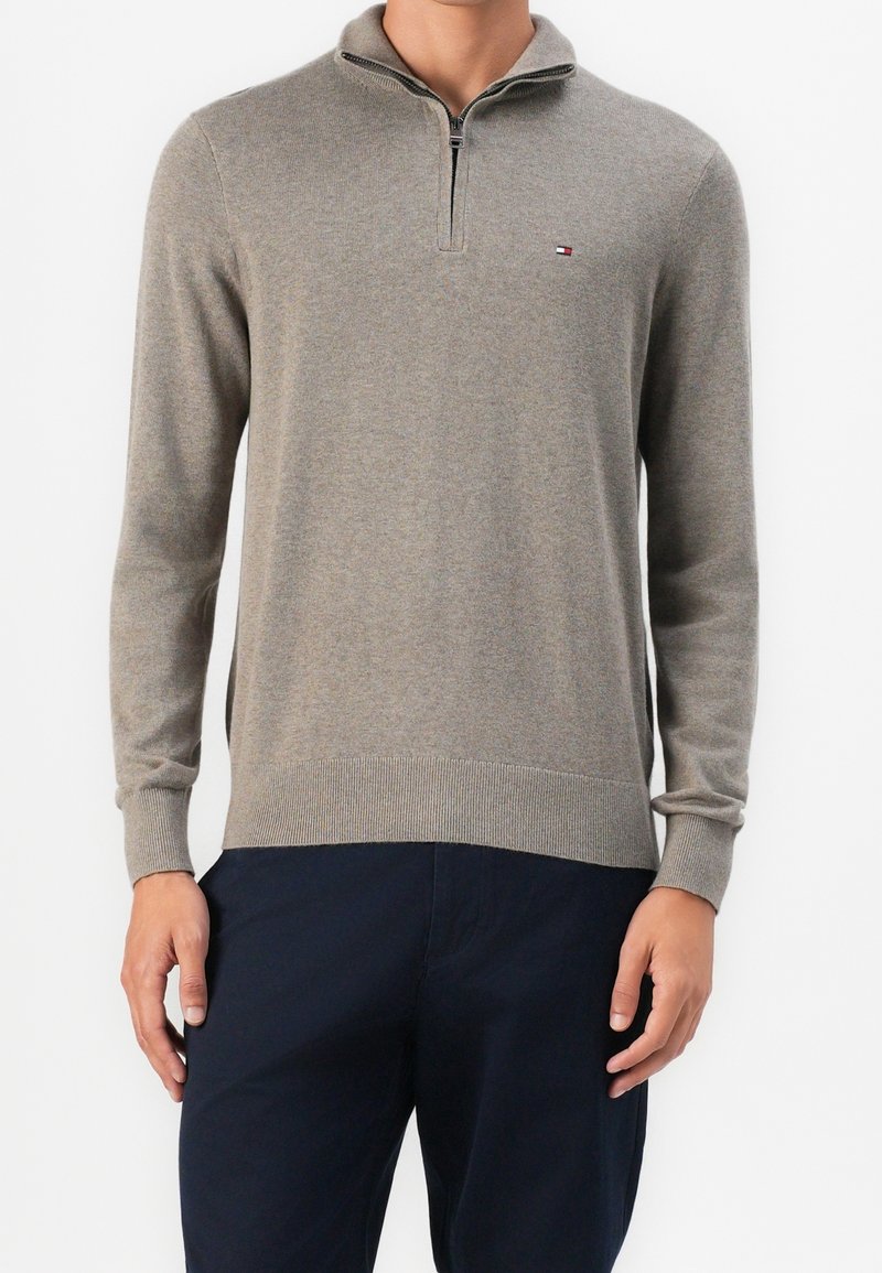 Homme portant un pull beige à manches longues avec fermeture quart de zip et petit logo sur la poitrine, et un pantalon bleu marine, debout sur fond blanc.