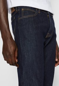 Dark blue denim jeans worn by a person with a white shirt, showing side pockets, belt loops, and metal rivets.