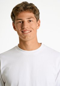 Vit långärmad T-shirt med rund halsringning på en person med kort brunt hår som ler mot en enkel ljus bakgrund. Mjukt, slätt tyg.