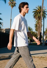 Mladić u bijeloj majici i sivim trapericama hoda na otvorenom, u pozadini su palme i vedro plavo nebo na sunčan dan.