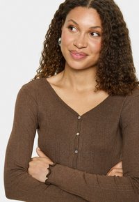 Vrouw met krullend haar, gekleed in een bruin geribd shirt met lange mouwen en knopen, met de armen gekruist, kijkt zijwaarts met een lichte glimlach.
