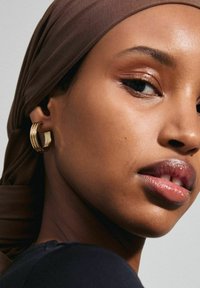 Pendientes de aro de oro con diseño en capas, llevados por una persona con un pañuelo en la cabeza de color marrón oscuro. La piel tiene un brillo natural y los labios parecen brillantes.