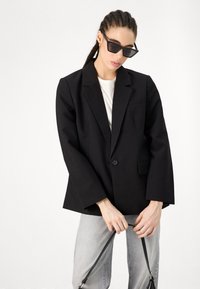 Ženska s spletenimi lasmi nosi mačje oči sončna očala, črno blazer, belo srajco, svetlo siva kavbojka, drži trak od črne torbice pred belo ozadje.